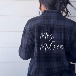 May include: A dark blue and black plaid button-down shirt featuring the white text "Mrs. McCrea" printed on the back. The upper left corner includes the text "thrifted + personalized!" in a simple black font against a light background.