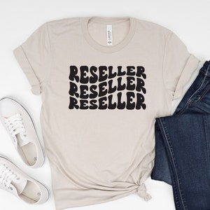 Puede incluir: Una camiseta beige con la palabra "RESELLER" impresa en negro, repetida tres veces en una fuente retro ondulada. La camiseta se combina con jeans azules y zapatillas blancas.