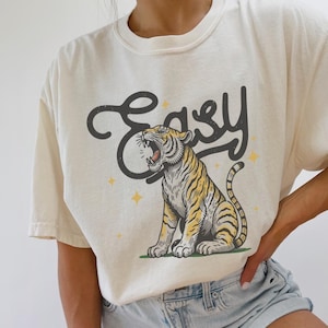 Puede incluir: Una camiseta blanca con un gr&aacute;fico de un tigre rugiente y la palabra "Easy" en una fuente estilizada. El tigre es amarillo y naranja con rayas negras. Hay peque&ntilde;as estrellas doradas alrededor del tigre.