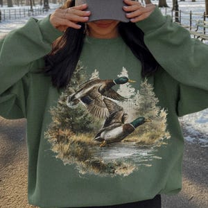 May include: An olive green sweatshirt featuring a graphic of two ducks in flight over a body of water, with a forest backdrop. The person is wearing a gray baseball cap with a white "NY" logo.
