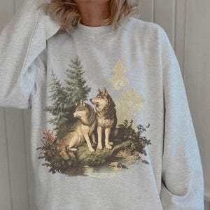 May include: A light grey sweatshirt with a detailed illustration of two wolves in a forest scene. The design includes trees, foliage, and a small body of water, creating a nature-themed design. The wolves are depicted with realistic fur and poses.