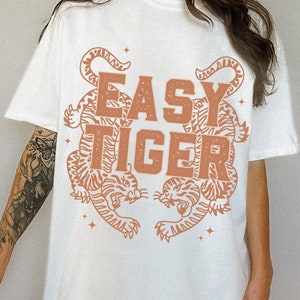Puede incluir: Camiseta blanca de gran tama&ntilde;o con un estampado gr&aacute;fico de dos tigres naranjas enfrentados con el texto "EASY TIGER" en el centro.