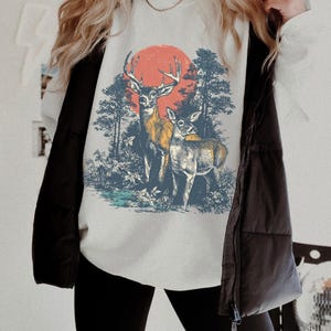 Op de afbeelding: Wit sweatshirt met een bossc&egrave;ne met twee herten en een rode zon. Het grafische ontwerp omvat bomen en gebladerte in blauwe en groene tinten. Onderdeel van een casual kledingcollectie.
