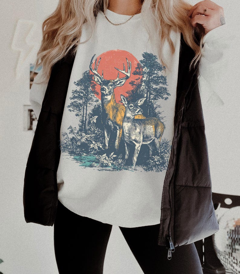 Op de afbeelding: Wit sweatshirt met een bossc&egrave;ne met twee herten en een rode zon. Het grafische ontwerp omvat bomen en gebladerte in blauwe en groene tinten. Onderdeel van een casual kledingcollectie.