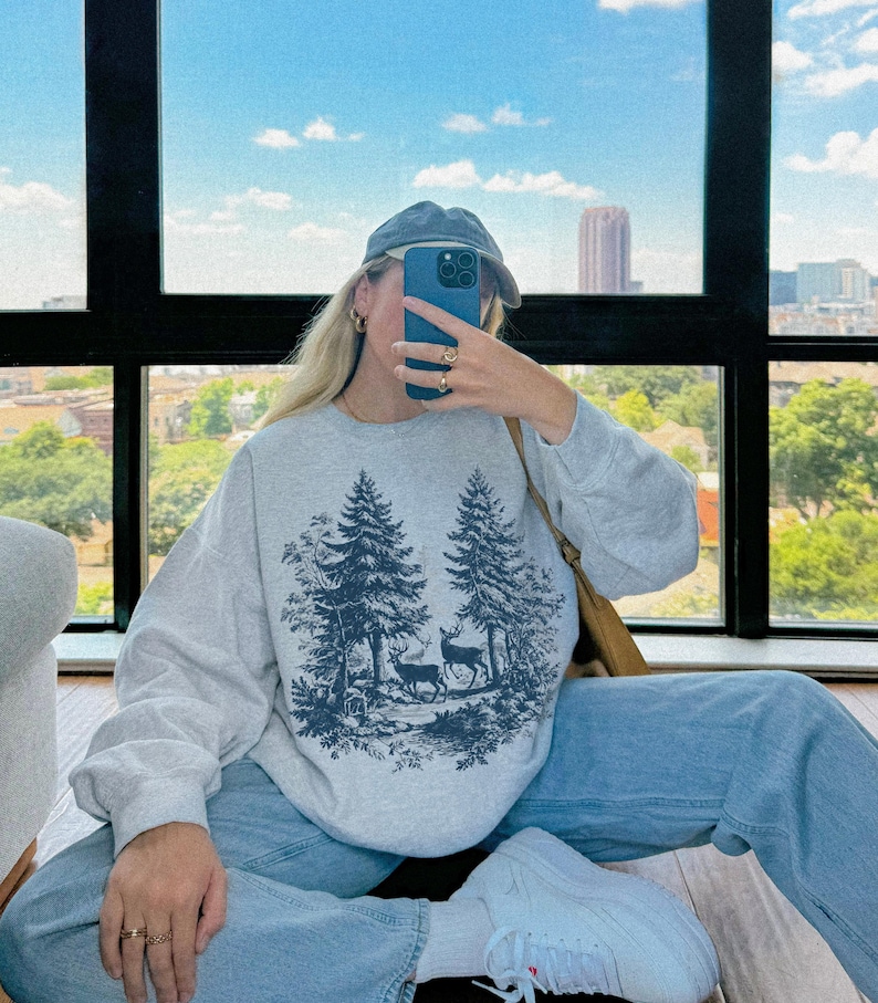 May include: A light gray sweatshirt featuring a forest scene with two deer and pine trees in a dark blue color. The sweatshirt is paired with light blue jeans and white sneakers. A blue cap is worn.