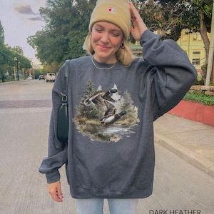 May include: Dark heather gray sweatshirt featuring a detailed graphic of two ducks in flight over a body of water, surrounded by trees. The text "DARK HEATHER" is printed at the bottom. The sweatshirt has long sleeves and a crew neck.