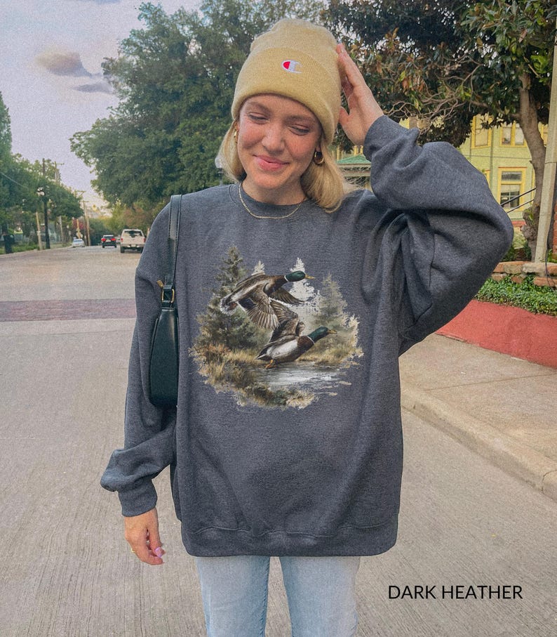 May include: Dark heather gray sweatshirt featuring a detailed graphic of two ducks in flight over a body of water, surrounded by trees. The text "DARK HEATHER" is printed at the bottom. The sweatshirt has long sleeves and a crew neck.