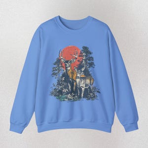 Op de afbeelding: Carolina blauwe sweatshirt met een afbeelding van twee herten in een bos en een rode zon. Het ontwerp is gedrukt op de voorkant van de sweatshirt met lange mouwen en ronde hals. De tekst "CAROLINA BLUE" staat onderaan.