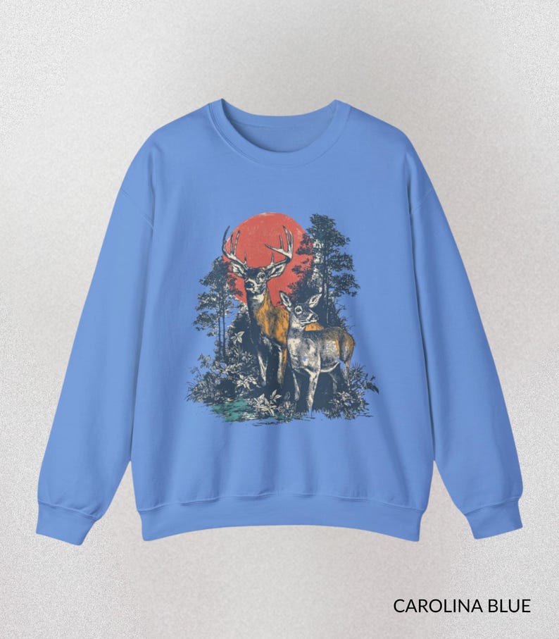 Op de afbeelding: Carolina blauwe sweatshirt met een afbeelding van twee herten in een bos en een rode zon. Het ontwerp is gedrukt op de voorkant van de sweatshirt met lange mouwen en ronde hals. De tekst "CAROLINA BLUE" staat onderaan.