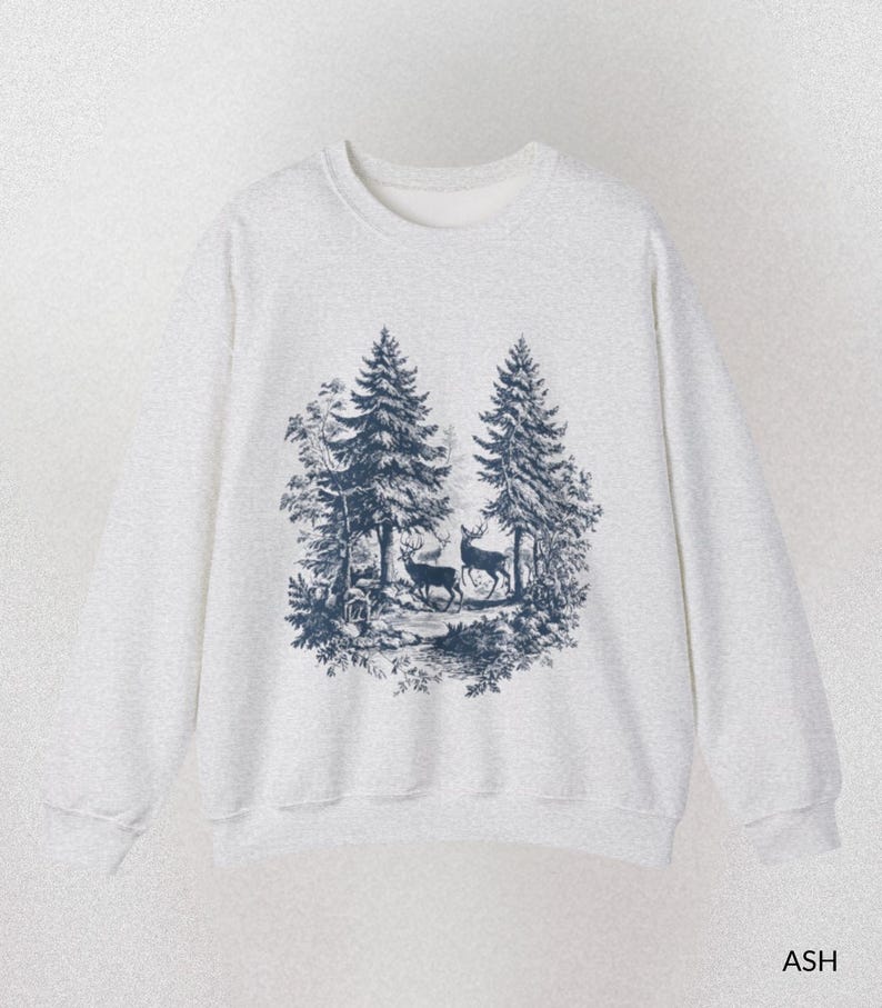 May include: A light gray sweatshirt with a crew neck. The front features a dark blue illustration of two deer in a forest scene with trees. The word "ASH" is printed in the bottom right corner.
