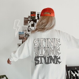 Peut inclure: Sweat-shirt blanc avec les mots "STINK STANK STUNK" imprim&eacute;s en superposition. La personne porte une casquette de camionneur orange. L'arri&egrave;re-plan comprend un mur blanc avec un &eacute;clair au n&eacute;on et un collage de photos.