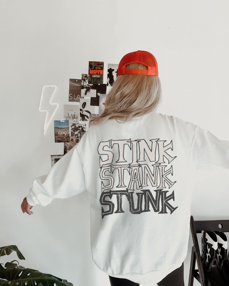 Peut inclure: Sweat-shirt blanc avec les mots "STINK STANK STUNK" imprim&eacute;s en superposition. La personne porte une casquette de camionneur orange. L'arri&egrave;re-plan comprend un mur blanc avec un &eacute;clair au n&eacute;on et un collage de photos.