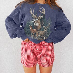 May include: A denim blue sweatshirt featuring a deer graphic with a forest backdrop. Paired with red and white checkered shorts. The word "DENIM" is printed at the bottom right corner of the image.