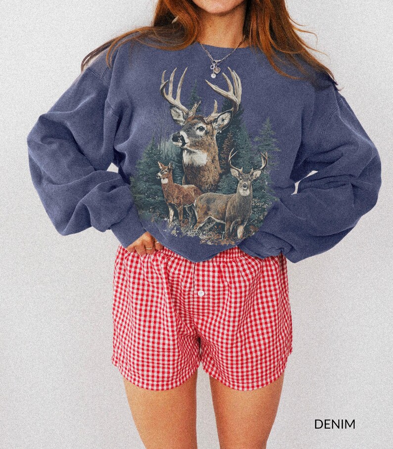 May include: A denim blue sweatshirt featuring a deer graphic with a forest backdrop. Paired with red and white checkered shorts. The word "DENIM" is printed at the bottom right corner of the image.