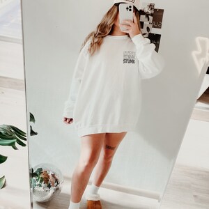 Peut inclure: Sweat-shirt blanc avec le texte "SHINE BLING STUNK" superpos&eacute;. Une casquette de baseball beige avec du texte est port&eacute;e. L'image est refl&eacute;t&eacute;e dans un miroir, montrant une boule disco et des pantoufles marron.