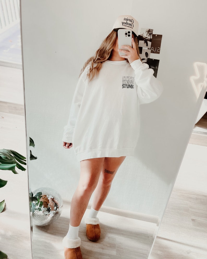 Peut inclure: Sweat-shirt blanc avec le texte "SHINE BLING STUNK" superpos&eacute;. Une casquette de baseball beige avec du texte est port&eacute;e. L'image est refl&eacute;t&eacute;e dans un miroir, montrant une boule disco et des pantoufles marron.