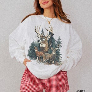 May include: White sweatshirt featuring a detailed illustration of deer in a forest setting. The graphic includes three deer with large antlers and evergreen trees. The shorts are red and white checkered.