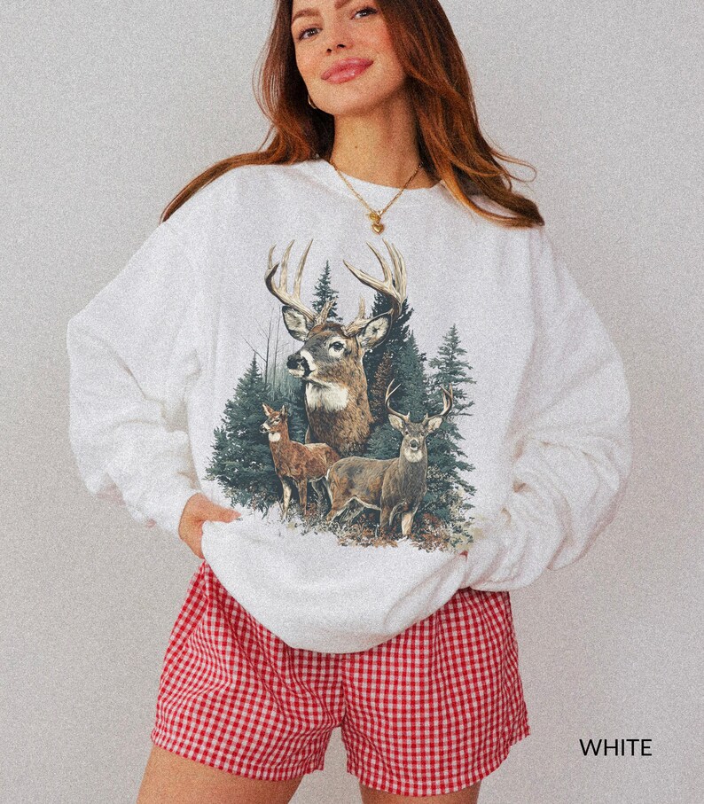 May include: White sweatshirt featuring a detailed illustration of deer in a forest setting. The graphic includes three deer with large antlers and evergreen trees. The shorts are red and white checkered.