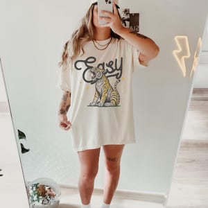 Puede incluir: Una camiseta blanca de gran tama&ntilde;o con un gr&aacute;fico de un tigre con la palabra "Easy" impresa encima. La modelo lleva una gorra de b&eacute;isbol con la palabra "Cruise" impresa en ella.
