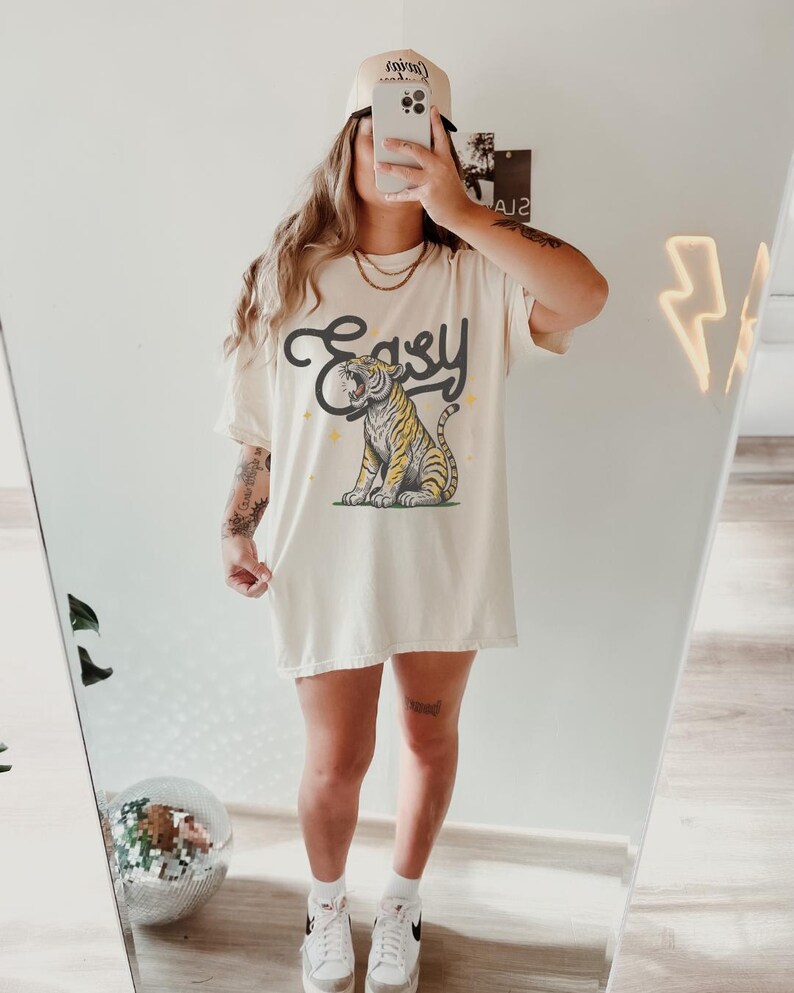 Puede incluir: Una camiseta blanca de gran tama&ntilde;o con un gr&aacute;fico de un tigre con la palabra "Easy" impresa encima. La modelo lleva una gorra de b&eacute;isbol con la palabra "Cruise" impresa en ella.