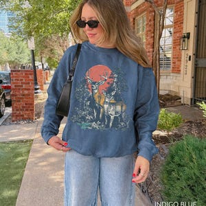 Op de afbeelding: Indigo blauwe sweatshirt met een hertenafbeelding, rode zon en bosachtergrond. De top met lange mouwen wordt gecombineerd met lichtblauwe jeans. De tekst "INDIGO BLUE" is rechtsonder in de afbeelding zichtbaar.