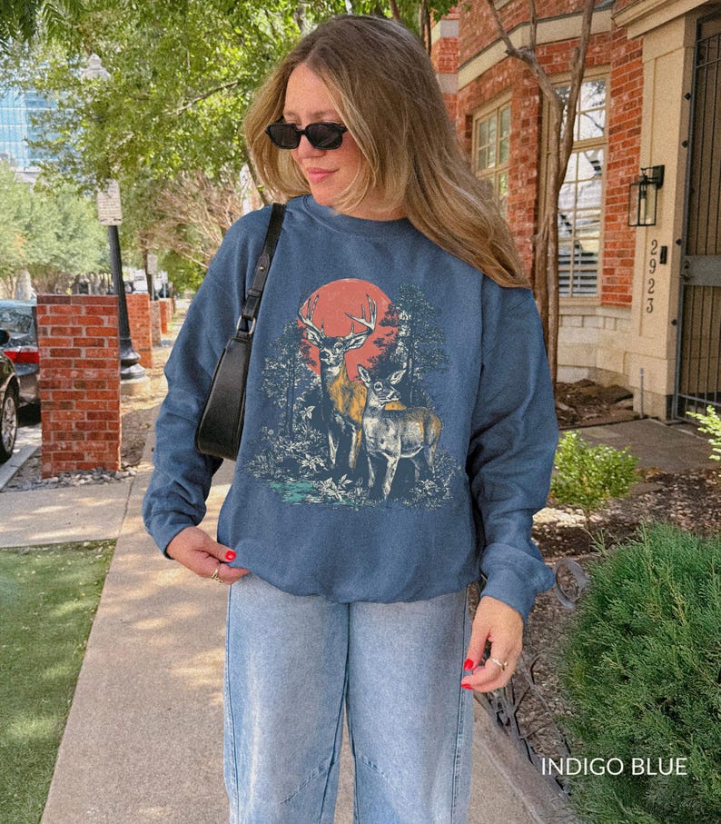 Op de afbeelding: Indigo blauwe sweatshirt met een hertenafbeelding, rode zon en bosachtergrond. De top met lange mouwen wordt gecombineerd met lichtblauwe jeans. De tekst "INDIGO BLUE" is rechtsonder in de afbeelding zichtbaar.