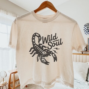 May include: A cream colored t-shirt with a black scorpion graphic and the text "Wild Soul" printed on the front.