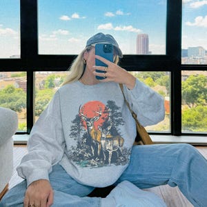 Op de afbeelding: Een lichtgrijs sweatshirt met een hertenafbeelding, een rode zon en bomen. Het sweatshirt wordt gedragen met lichtblauwe jeans en witte sneakers. Het woord "ASH" is rechtsonder zichtbaar.