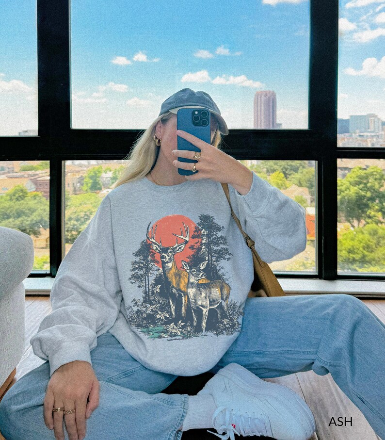 Op de afbeelding: Een lichtgrijs sweatshirt met een hertenafbeelding, een rode zon en bomen. Het sweatshirt wordt gedragen met lichtblauwe jeans en witte sneakers. Het woord "ASH" is rechtsonder zichtbaar.