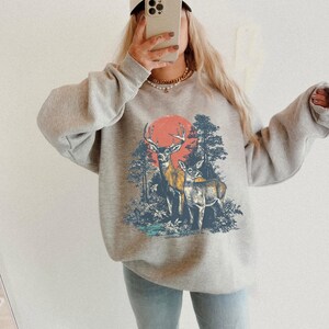 Op de afbeelding: Sportgrijs sweatshirt met een afbeelding van twee herten in een bos en een rode zon. De sweatshirt heeft lange mouwen en een ronde hals. Een beige baseballcap met het woord "KARMA" is zichtbaar. De tekst "SPORT GREY" staat onderaan.