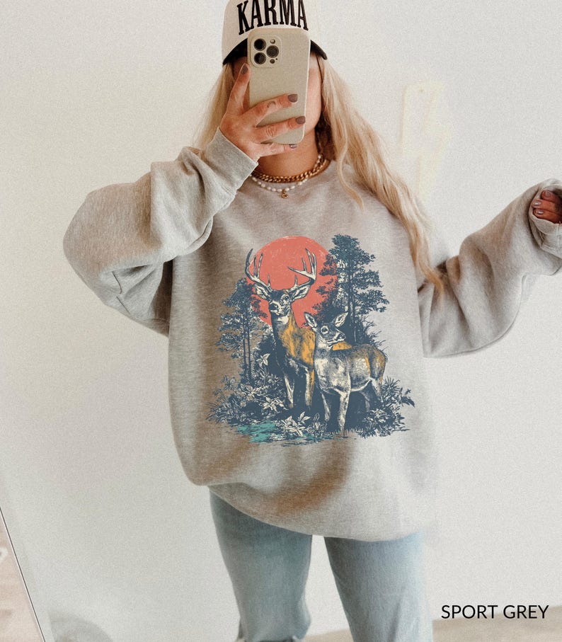 Op de afbeelding: Sportgrijs sweatshirt met een afbeelding van twee herten in een bos en een rode zon. De sweatshirt heeft lange mouwen en een ronde hals. Een beige baseballcap met het woord "KARMA" is zichtbaar. De tekst "SPORT GREY" staat onderaan.