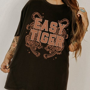 Puede incluir: Camiseta negra de gran tama&ntilde;o con un gr&aacute;fico marr&oacute;n de dos tigres y el texto "Easy Tiger".