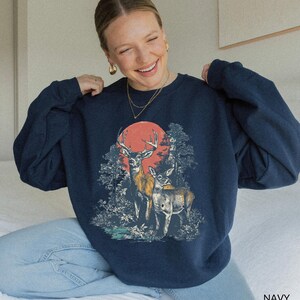 Op de afbeelding: Marineblauwe sweatshirt met een afbeelding van twee herten in een bos met een rode zon op de achtergrond. De sweatshirt heeft een ronde hals en lange mouwen. Het woord "NAVY" staat rechtsonder.