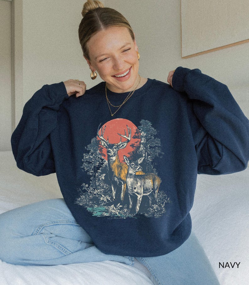 Op de afbeelding: Marineblauwe sweatshirt met een afbeelding van twee herten in een bos met een rode zon op de achtergrond. De sweatshirt heeft een ronde hals en lange mouwen. Het woord "NAVY" staat rechtsonder.