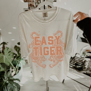 Puede incluir: Una camiseta blanca con un gr&aacute;fico en color melocot&oacute;n de dos tigres enfrentados. El texto "EASY TIGER" est&aacute; impreso en letras de color melocot&oacute;n sobre los tigres.