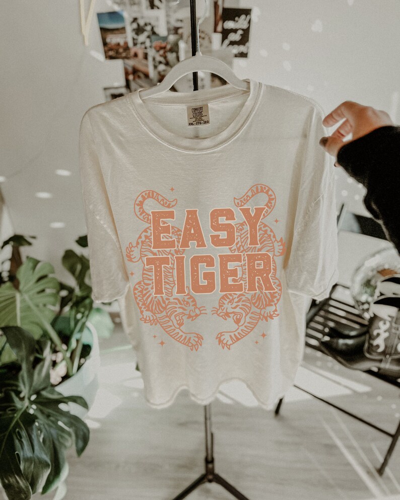 Puede incluir: Una camiseta blanca con un gr&aacute;fico en color melocot&oacute;n de dos tigres enfrentados. El texto "EASY TIGER" est&aacute; impreso en letras de color melocot&oacute;n sobre los tigres.