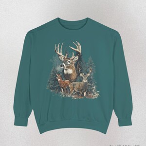 May include: Teal sweatshirt featuring a detailed illustration of three deer in a forest setting. The design includes a large buck with prominent antlers and two smaller deer. The text "BLUE SPRUCE" is printed at the bottom.