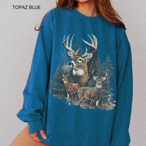 May include: Topaz blue sweatshirt featuring a detailed graphic of three deer in a forest setting. The design includes a large buck with prominent antlers, and the text "TOPAZ BLUE" is printed at the top.