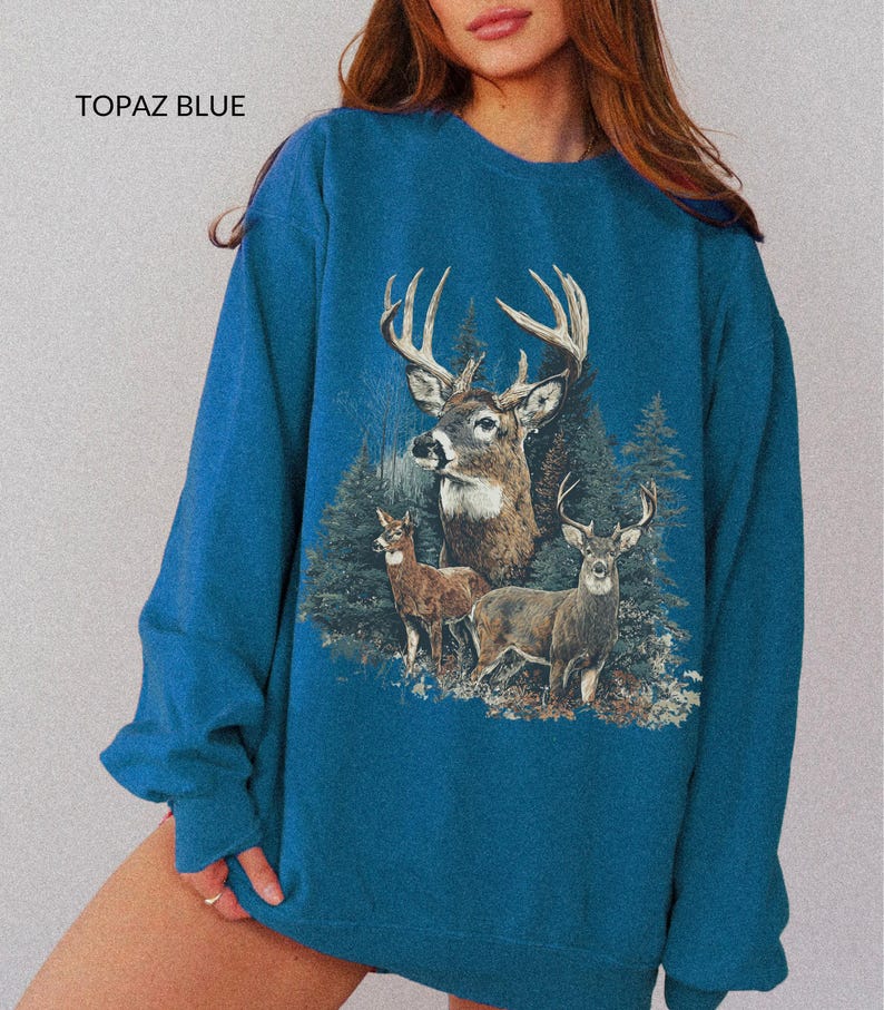 May include: Topaz blue sweatshirt featuring a detailed graphic of three deer in a forest setting. The design includes a large buck with prominent antlers, and the text "TOPAZ BLUE" is printed at the top.