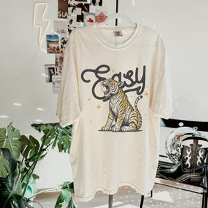 Puede incluir: Una camiseta de color crema con un gr&aacute;fico de un tigre rugiendo con la palabra "Easy" encima. El tigre es amarillo y negro sobre un fondo blanco.