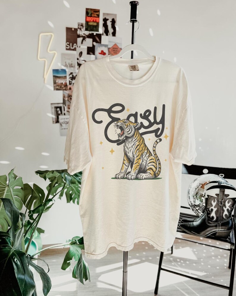 Puede incluir: Una camiseta de color crema con un gr&aacute;fico de un tigre rugiendo con la palabra "Easy" encima. El tigre es amarillo y negro sobre un fondo blanco.