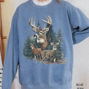 May include: A blue jean-colored sweatshirt featuring a detailed graphic of three deer in a forest setting. The design includes a large buck with prominent antlers and two smaller deer. The text "BLUE JEAN" is printed at the bottom right.