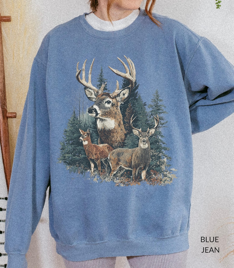 May include: A blue jean-colored sweatshirt featuring a detailed graphic of three deer in a forest setting. The design includes a large buck with prominent antlers and two smaller deer. The text "BLUE JEAN" is printed at the bottom right.