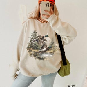 May include: Sand-colored sweatshirt featuring a detailed graphic of two ducks in flight over a body of water, surrounded by trees. The sweatshirt has a crew neck and long sleeves. The word "SAND" is printed in the bottom right corner.