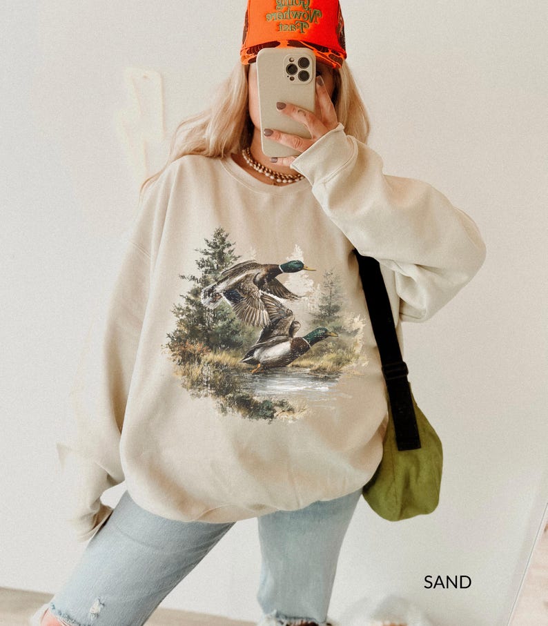 May include: Sand-colored sweatshirt featuring a detailed graphic of two ducks in flight over a body of water, surrounded by trees. The sweatshirt has a crew neck and long sleeves. The word "SAND" is printed in the bottom right corner.