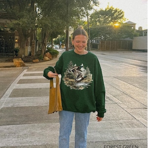 May include: Forest green sweatshirt featuring a nature scene with ducks. The sweatshirt has a crew neck and long sleeves. The text "FOREST GREEN" is printed at the bottom of the image. The person is wearing blue jeans and holding a brown tote bag.