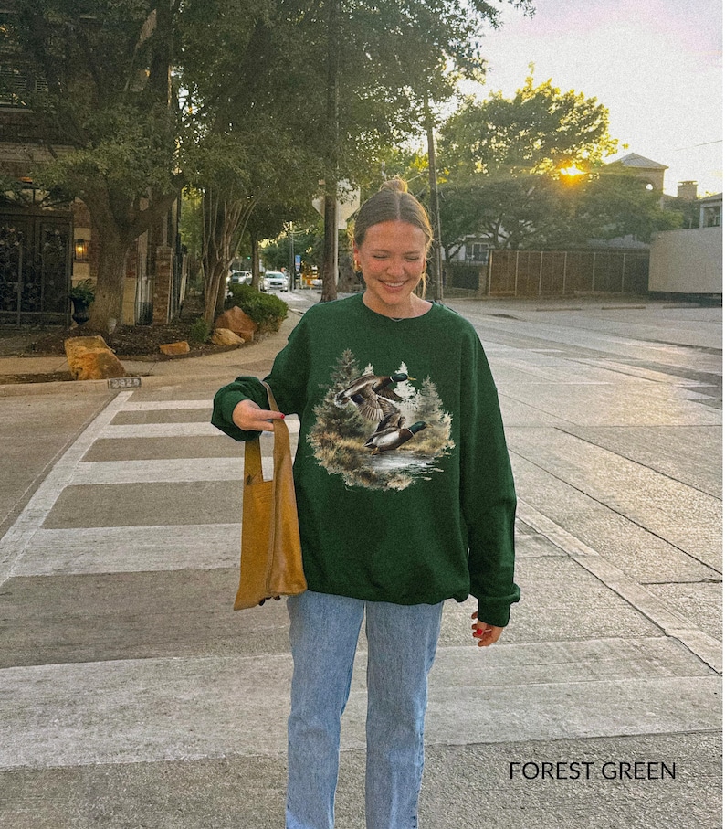 May include: Forest green sweatshirt featuring a nature scene with ducks. The sweatshirt has a crew neck and long sleeves. The text "FOREST GREEN" is printed at the bottom of the image. The person is wearing blue jeans and holding a brown tote bag.