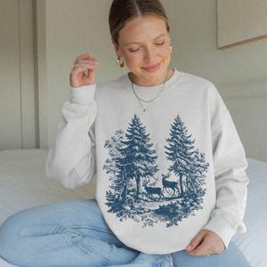 May include: A cream-colored sweatshirt featuring a blue forest scene with two deer and tall pine trees. The graphic design is centered on the front of the long-sleeved crew neck sweatshirt. The sweatshirt is made of a soft material.