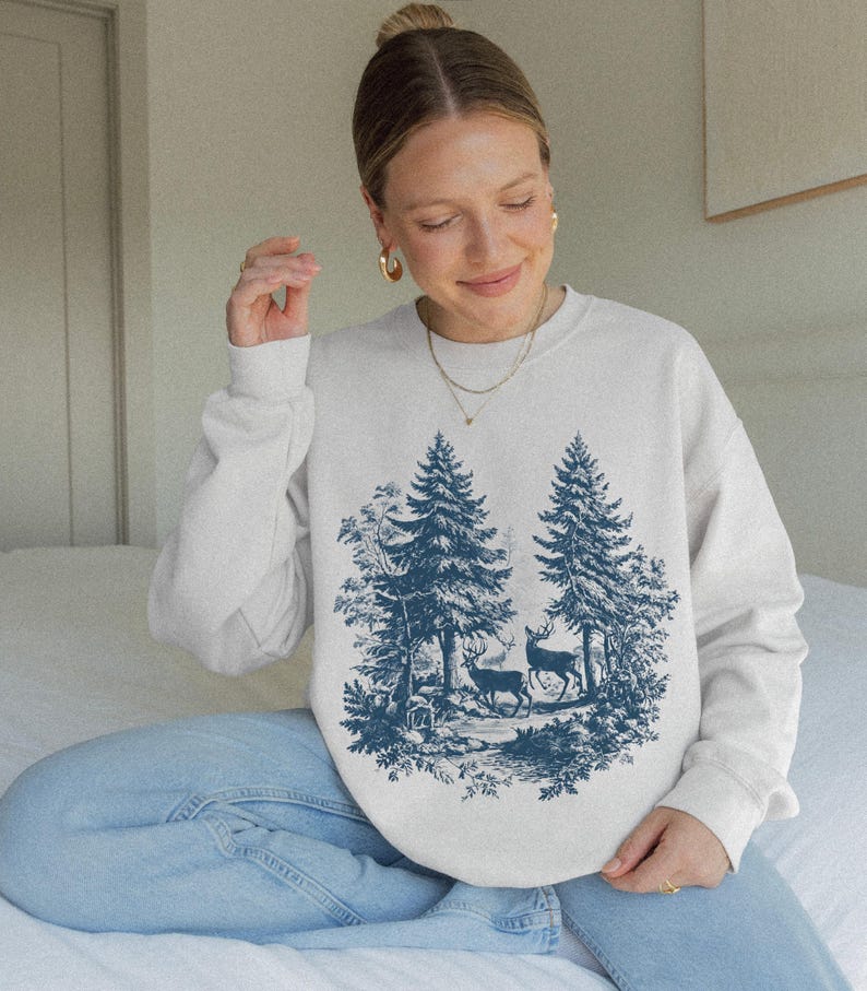 May include: A cream-colored sweatshirt featuring a blue forest scene with two deer and tall pine trees. The graphic design is centered on the front of the long-sleeved crew neck sweatshirt. The sweatshirt is made of a soft material.