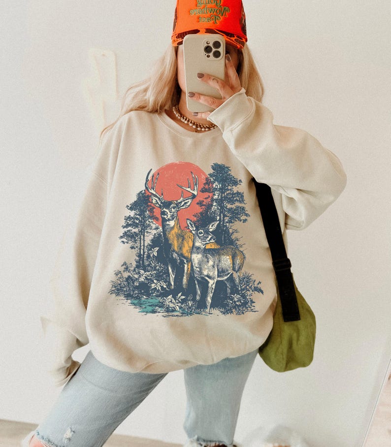 Op de afbeelding: Cr&egrave;mekleurige sweatshirt met een afbeelding van twee herten in een bos met een rode zon op de achtergrond. Het ontwerp is in tinten blauw, geel en bruin. De persoon draagt een feloranje hoed.